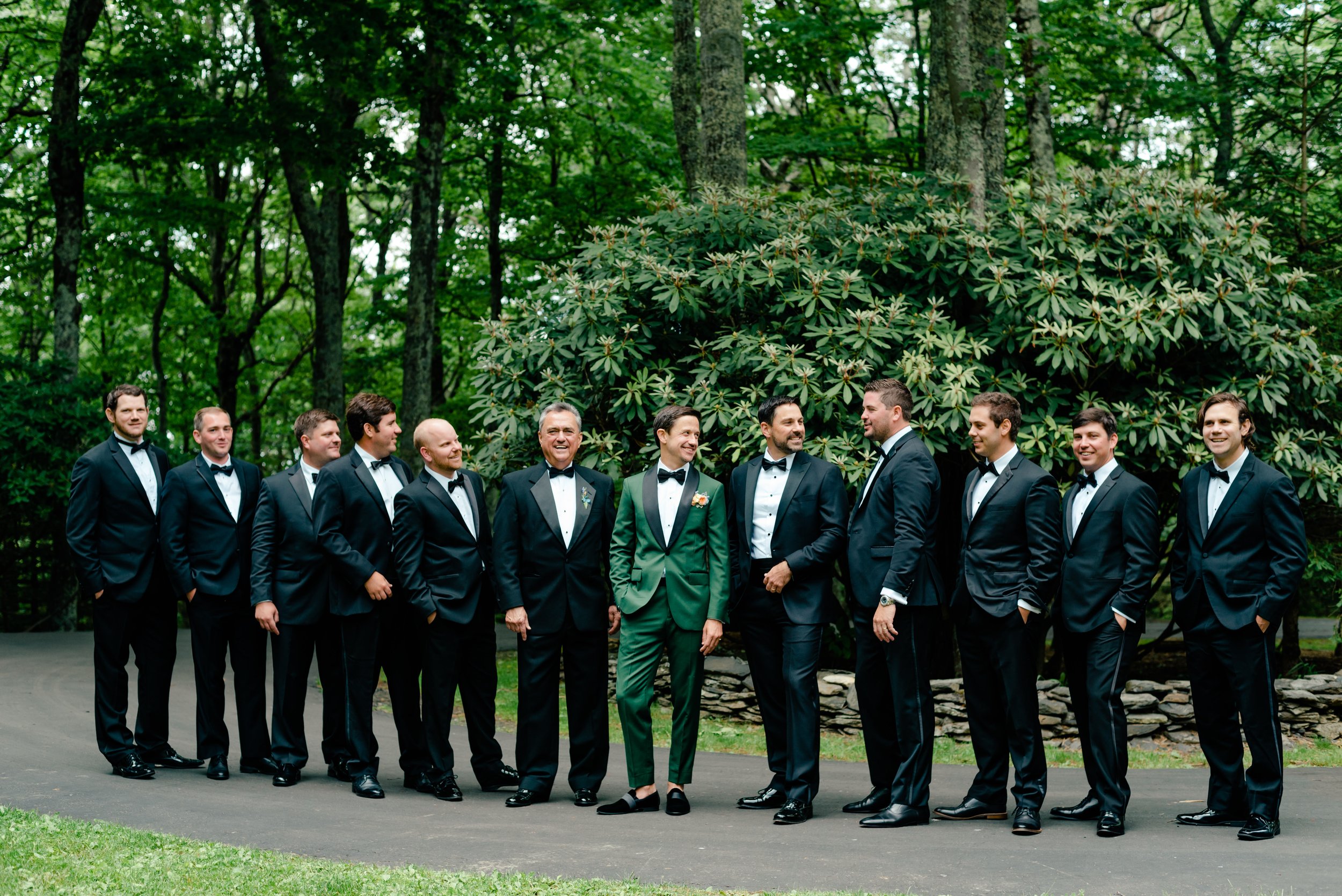 Groom in a green suit posing with groomsmen in black tuxedos for a group portrait outdoors
