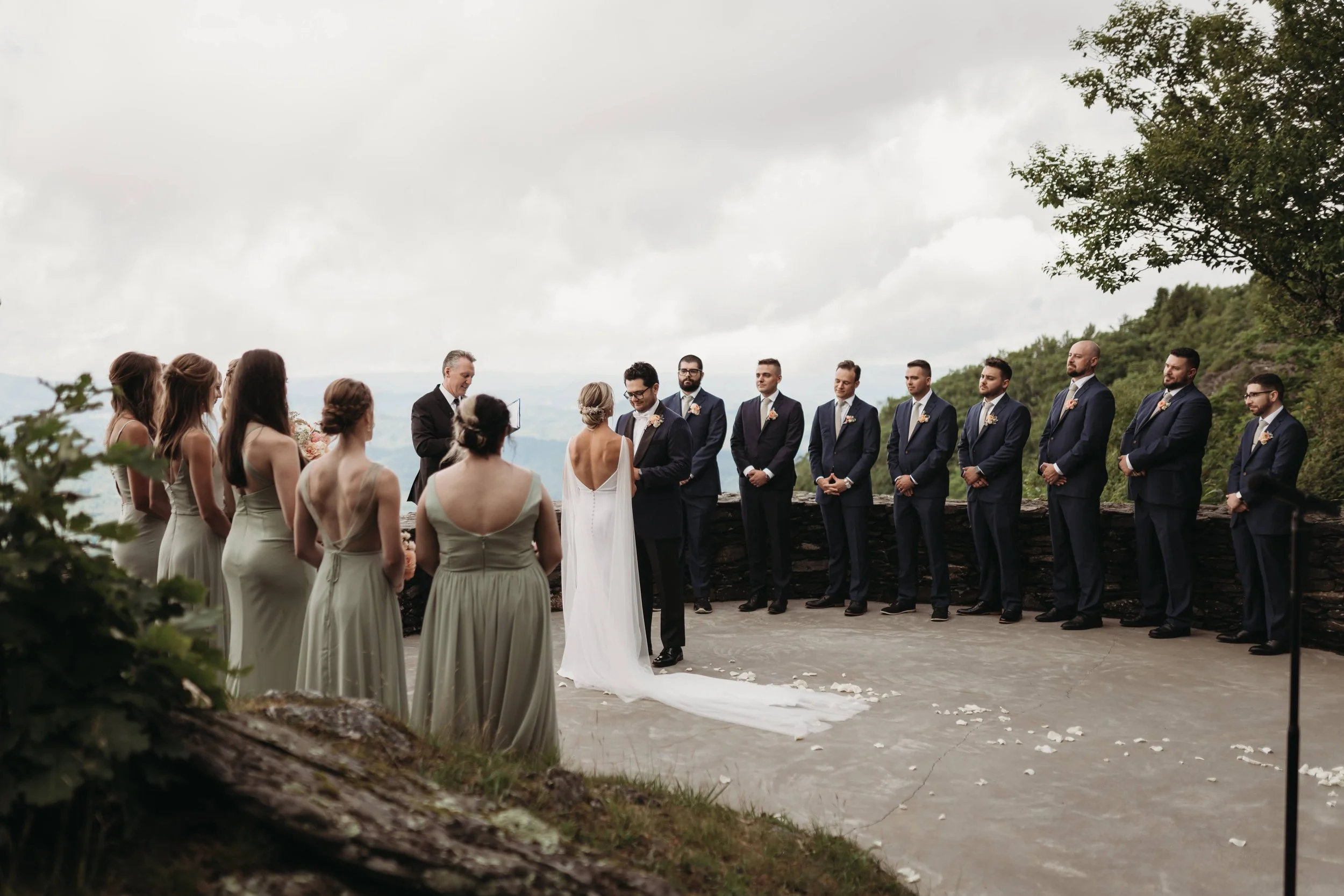 Outdoor mountain-view wedding ceremony with the couple at the altar, bridesmaids in sage dresses, and groomsmen in dark suits.