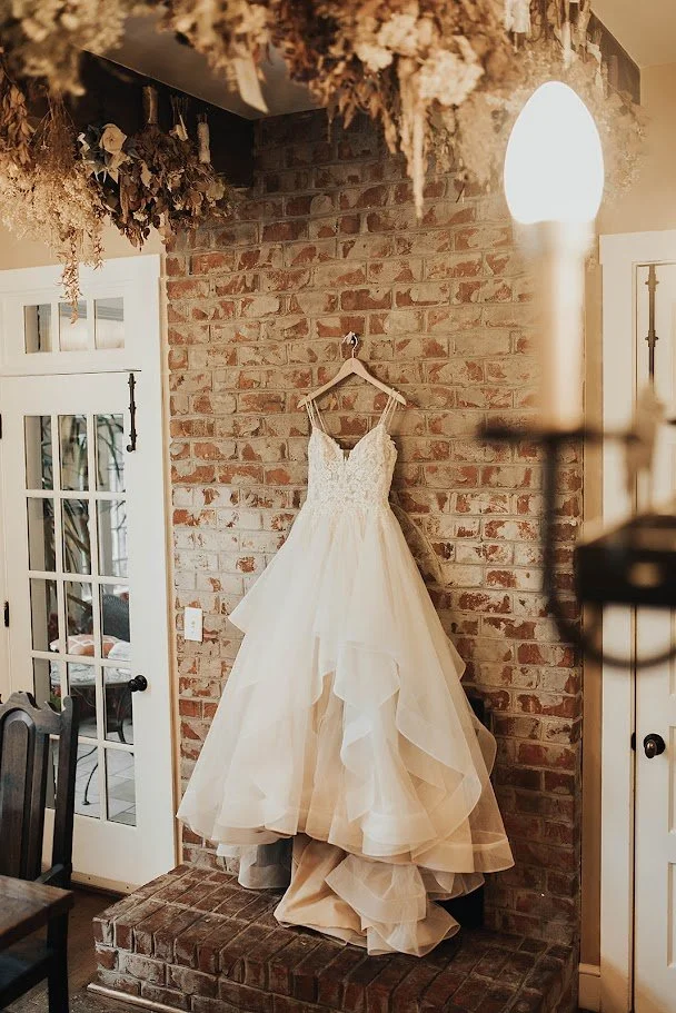 Wedding dress hanging on a brick wall in a bridal suite with dried florals overhead.