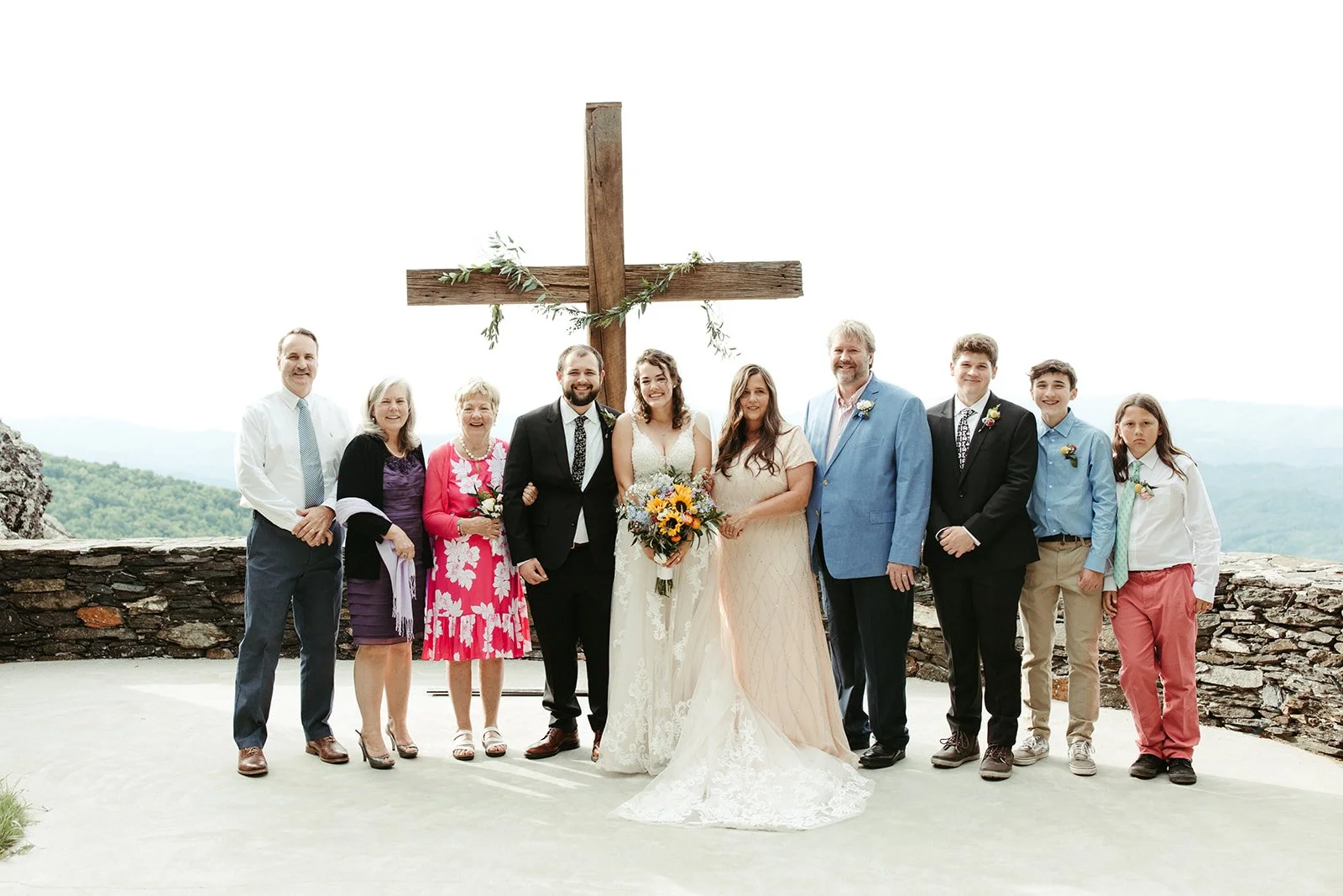 Bride and groom posing with family beneath a wooden cross at a mountain overlook wedding ceremony.