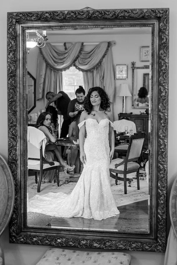 Black-and-white mirror portrait of the bride in an off-the-shoulder wedding gown while getting ready