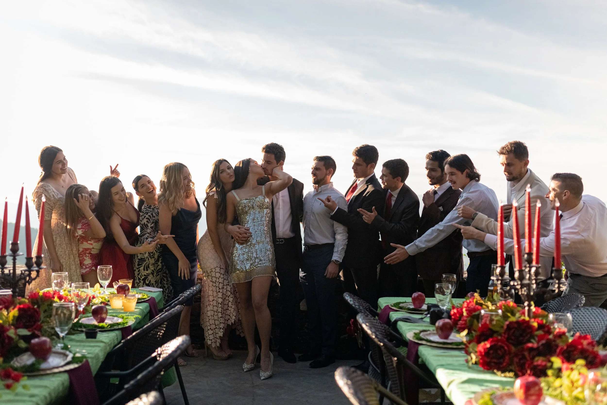 Guests posing together at an outdoor rehearsal dinner with colorful tables, red taper candles, and mountain views.