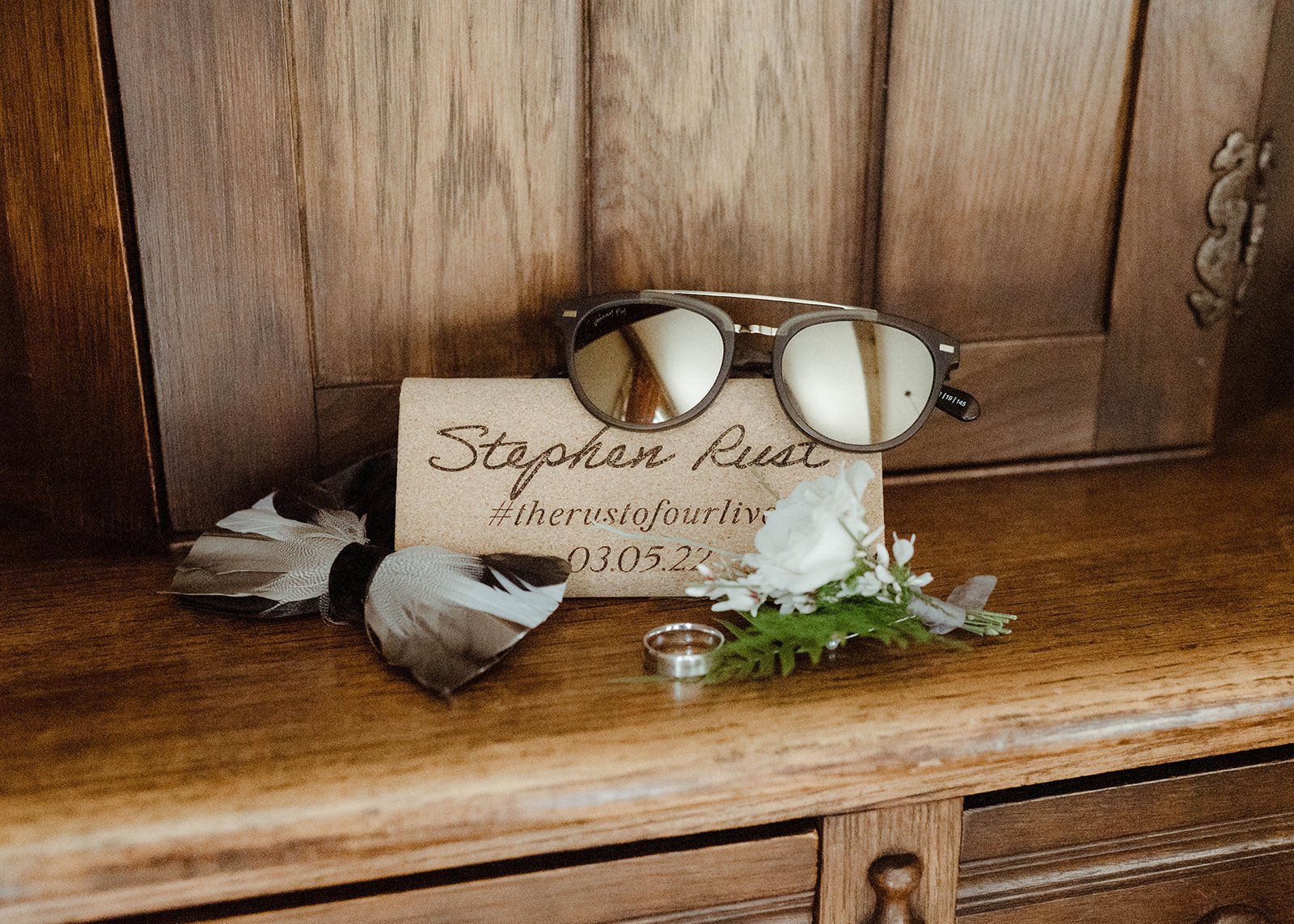 Groom detail flat lay with sunglasses, wedding ring, feathers, greenery, and personalized wood card.