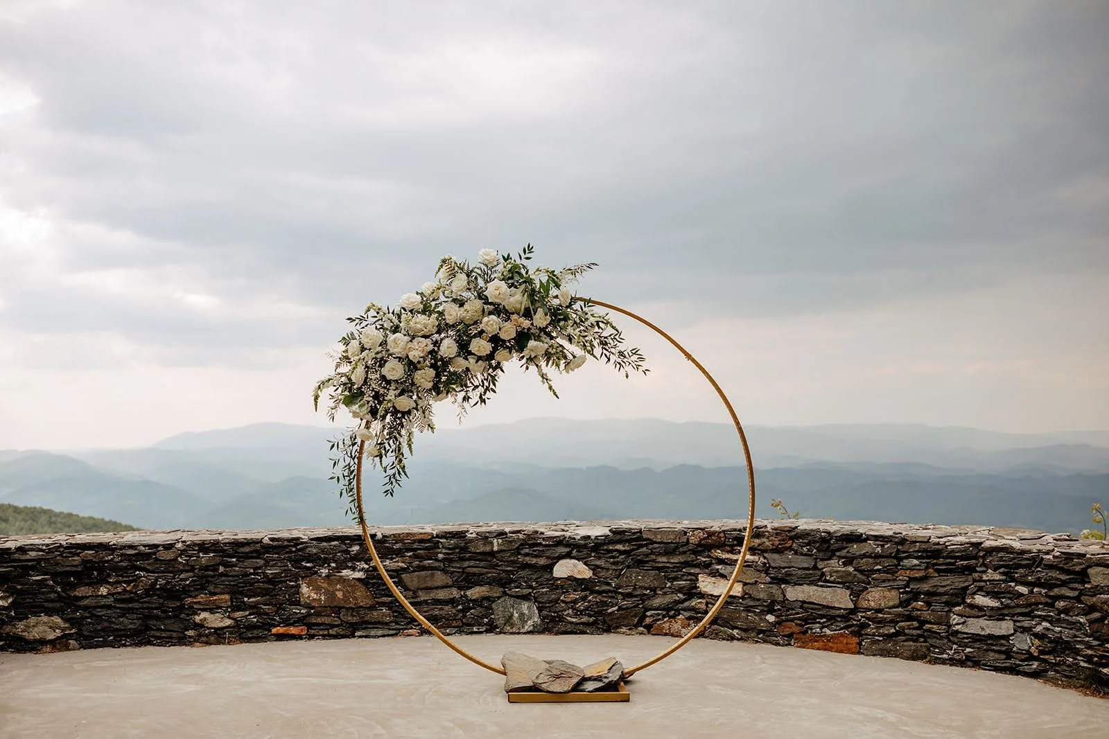 Circular wedding ceremony arch with white flowers and greenery on a stone terrace with mountain views