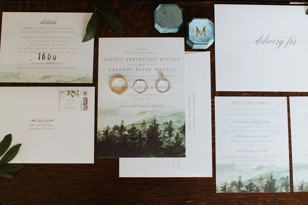 Wedding invitation suite flat lay with a mountain illustration and blue velvet ring boxes on a wooden surface.