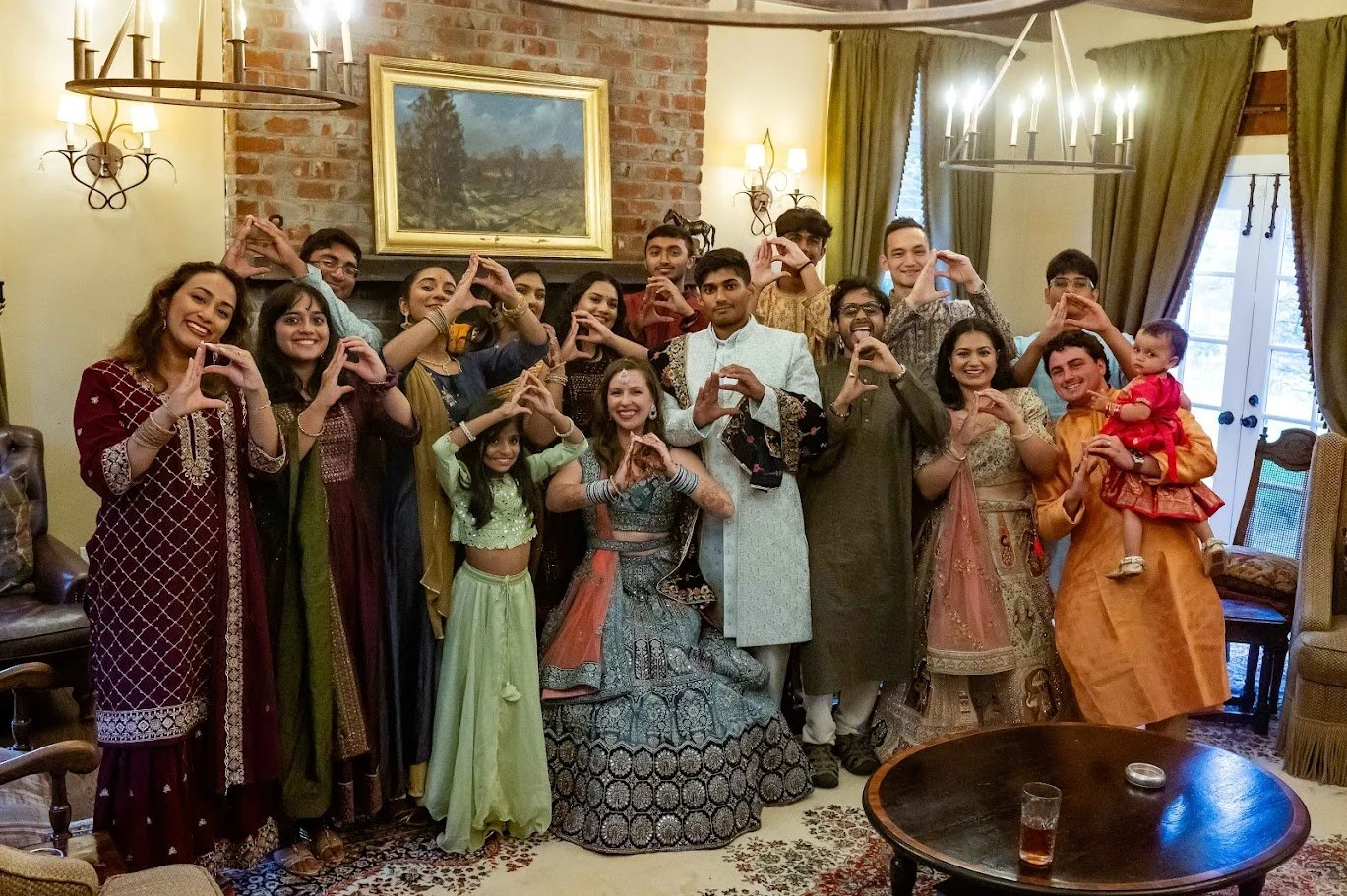 Guests indoors pose around the couple, many making heart shapes with their hands in a warmly lit room.