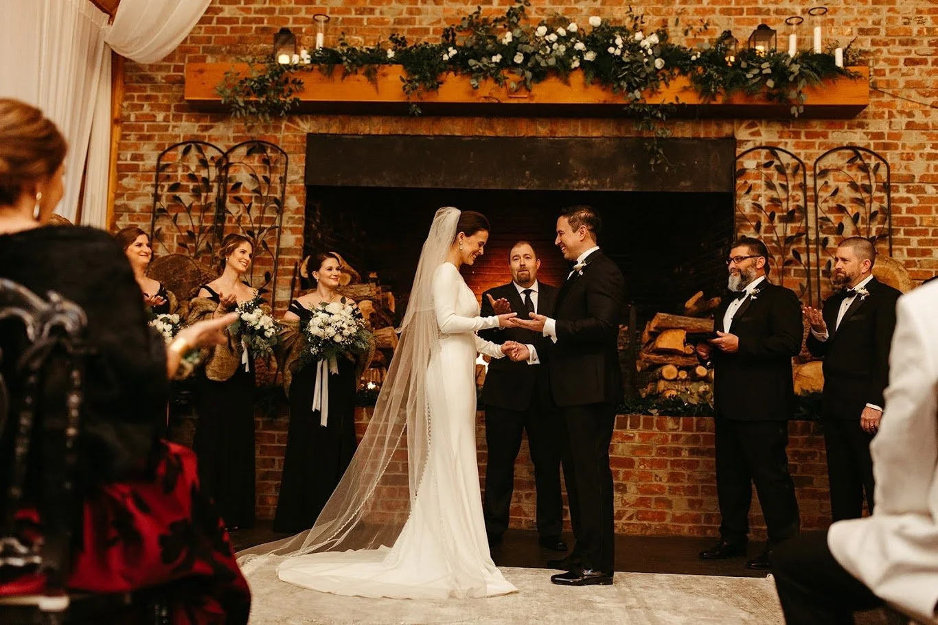 Wide view of an indoor wedding ceremony in front of a fireplace with florals and guests standing nearby.
