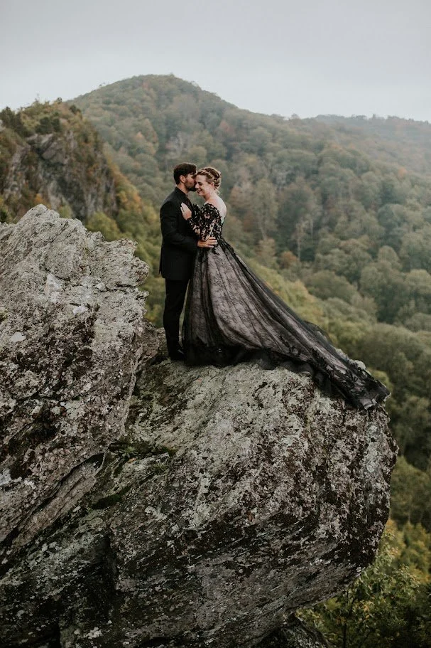 Bride and groom embracing on a rocky cliff with mountains behind them, bride wearing a flowing black gown.
