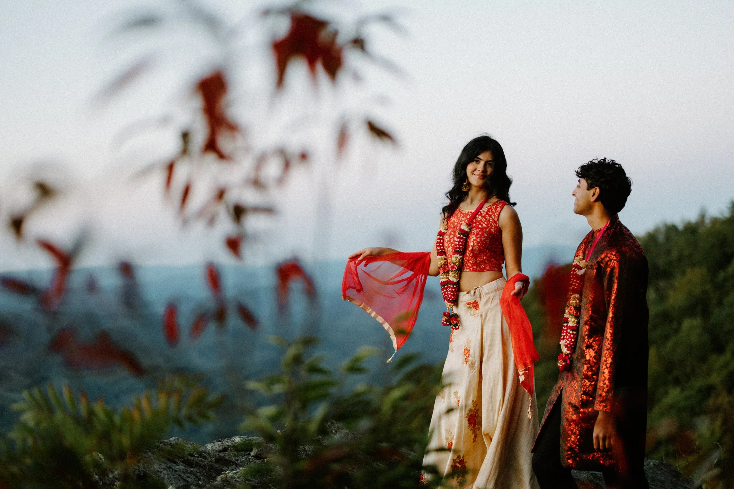 Couple in red and cream South Asian attire stands on an overlook with mountains behind, framed by soft foreground leaves.
