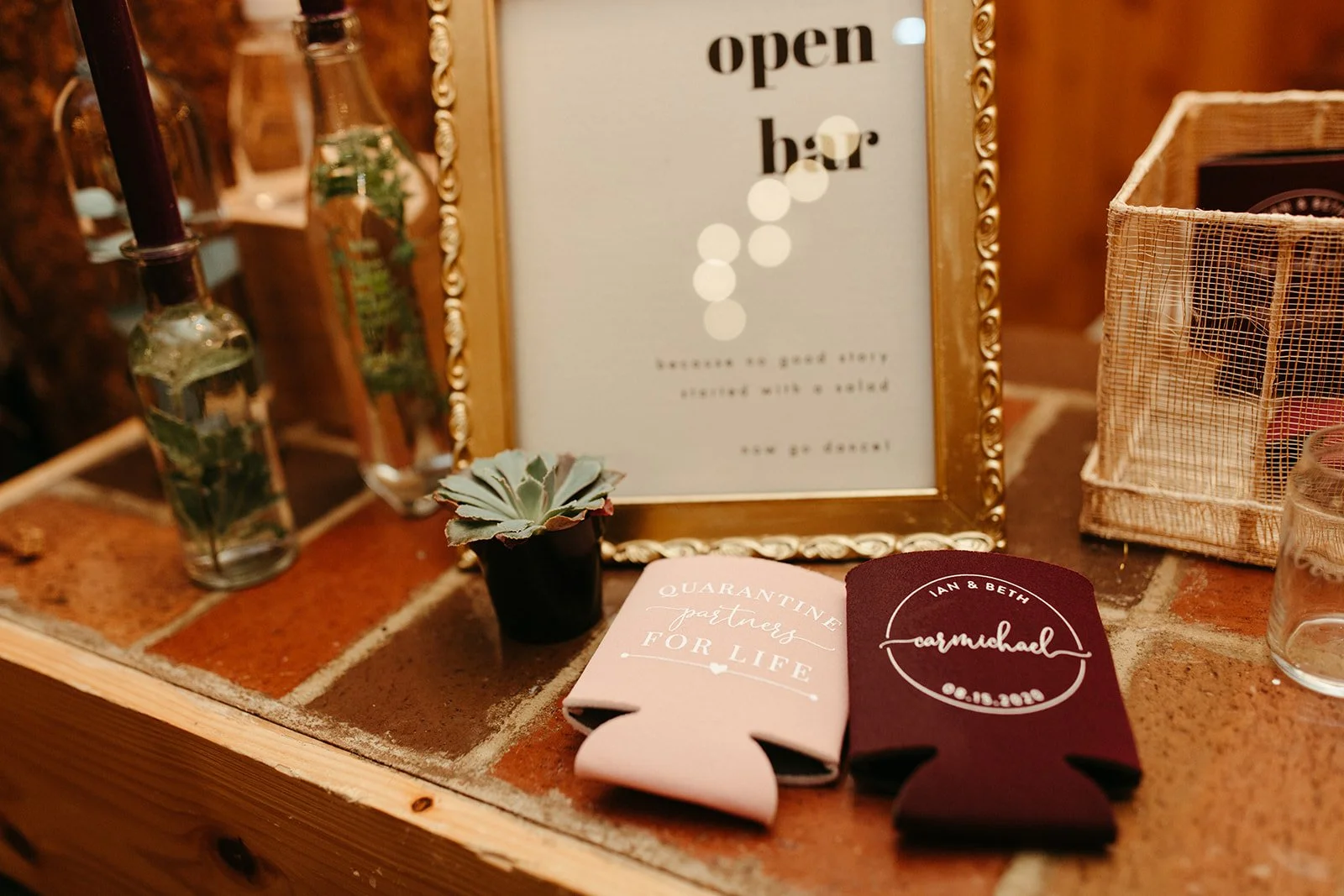 Wedding reception detail with an open bar sign, framed decor, and custom drink koozies on a brick ledge