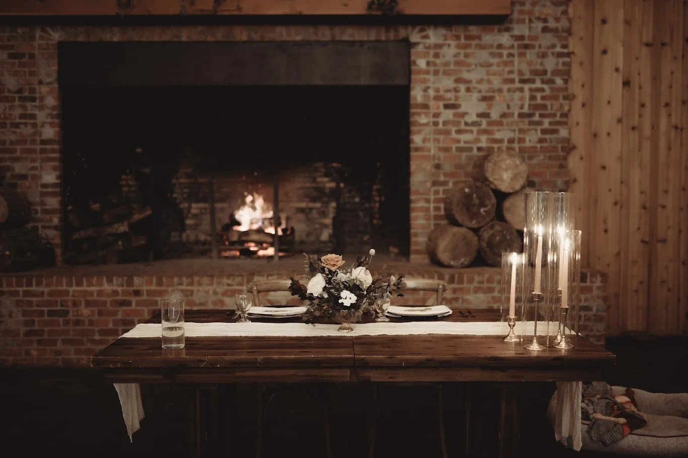Rustic sweetheart table set in front of a lit brick fireplace with candles and floral centerpiece