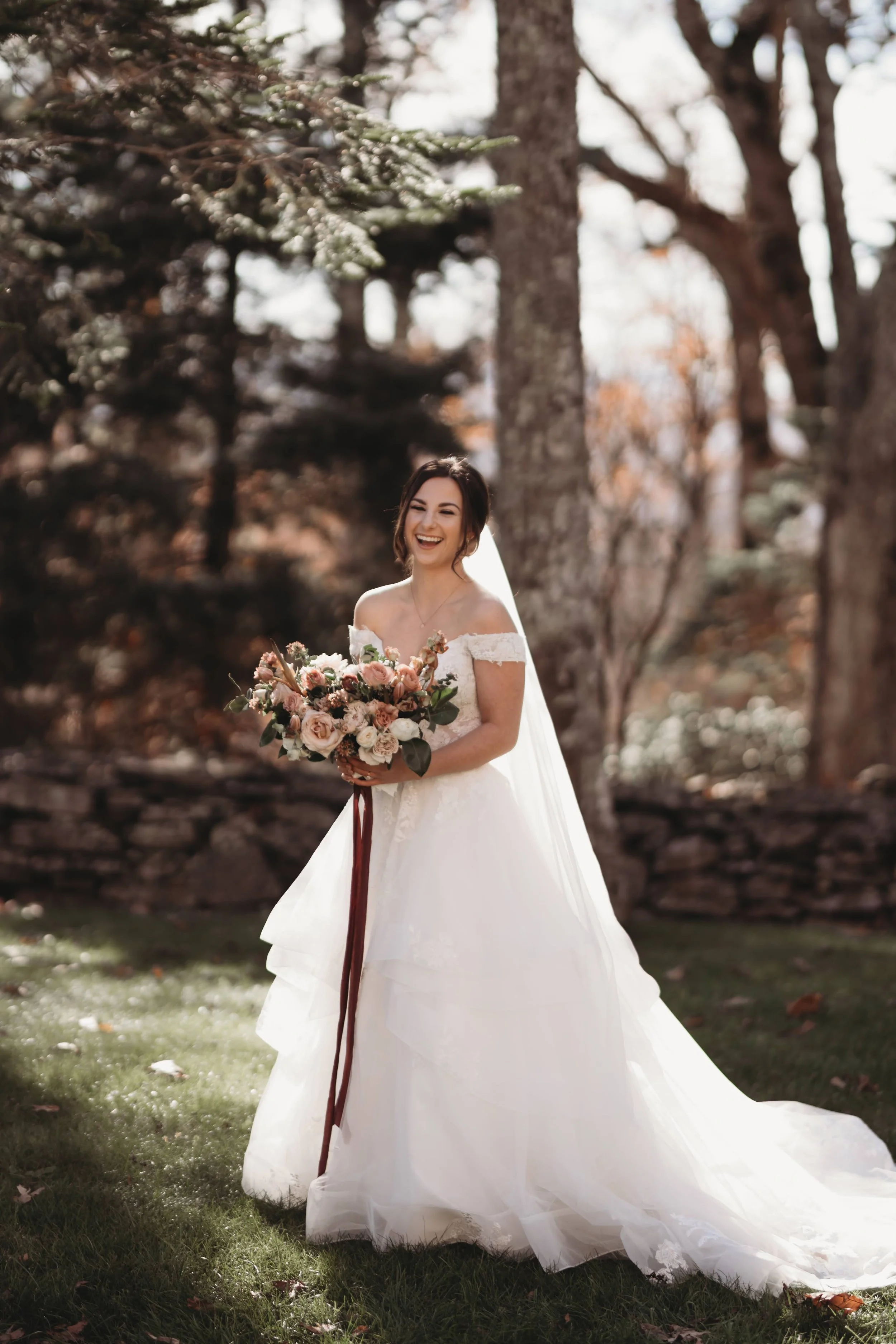 Bride laughing during outdoor portraits, holding a bouquet with a long veil behind her.