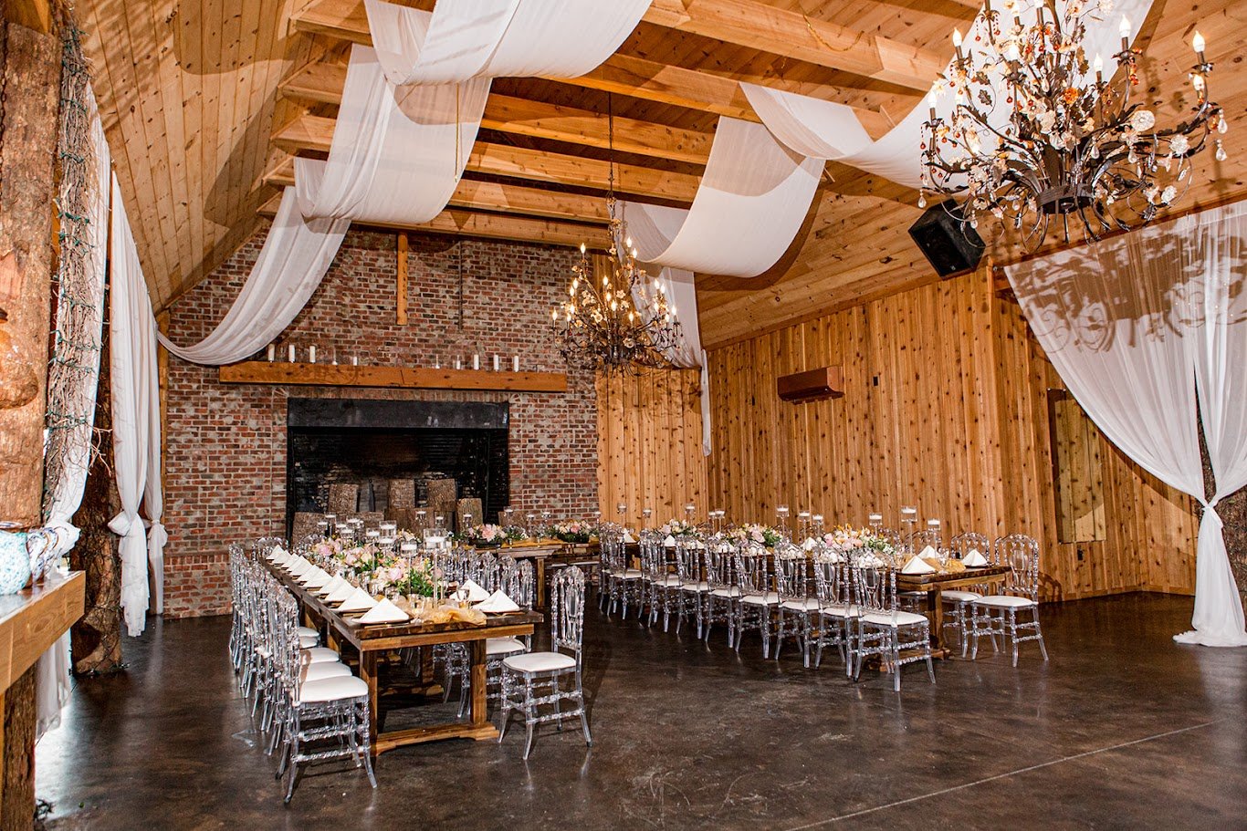 Wide view of a rustic barn wedding reception with long tables, clear chairs, chandeliers, and ceiling draping