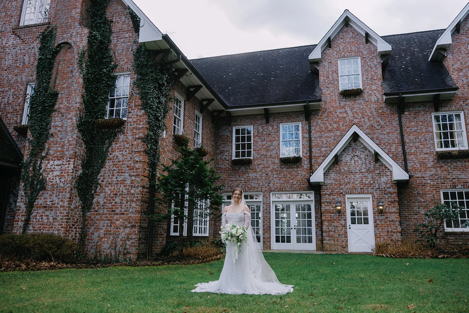 Bride standing on the lawn outside an ivy-covered brick estate wedding venue while holding a white bouquet.