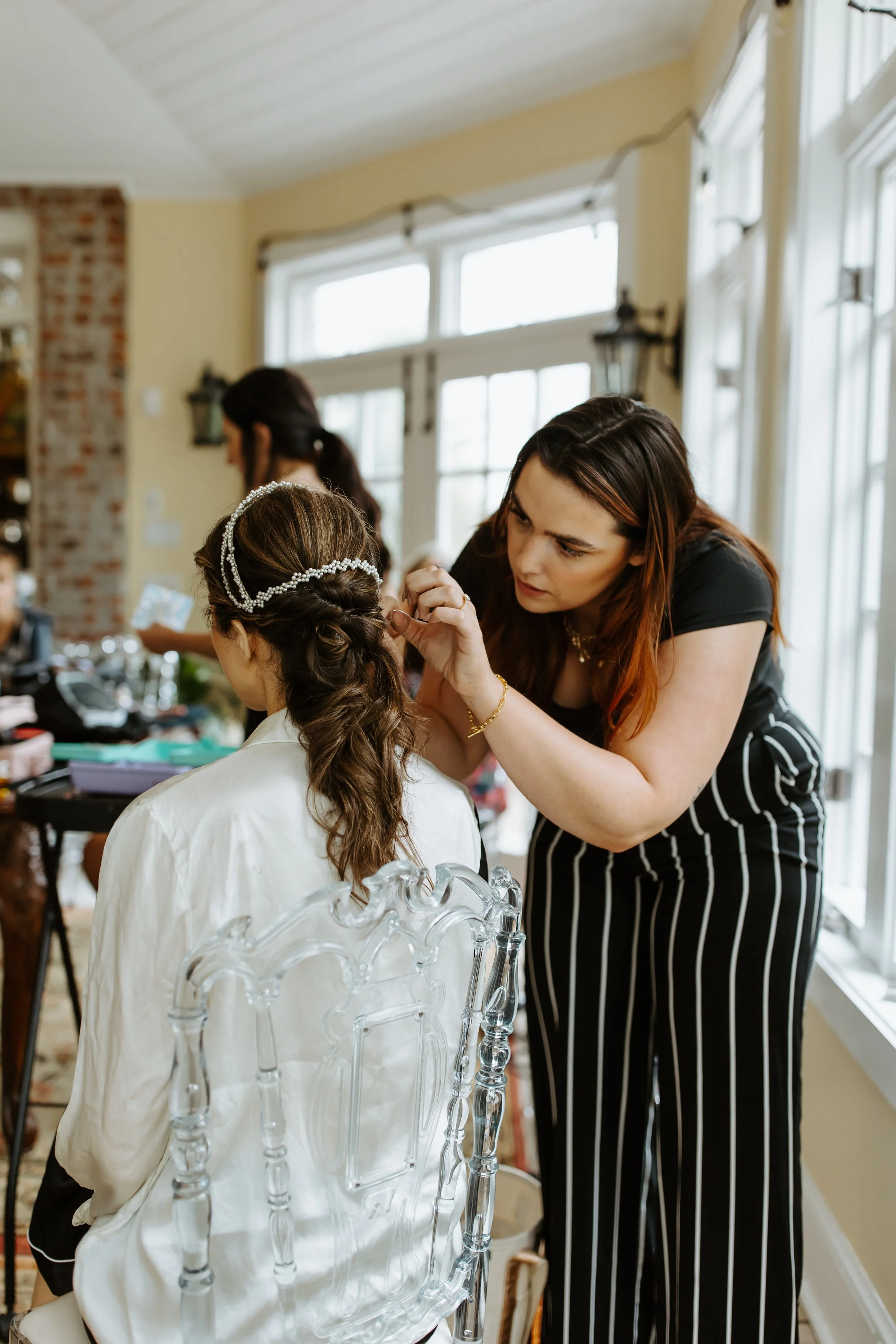 Hair stylist braiding the bride’s hair during getting ready in a bright room.