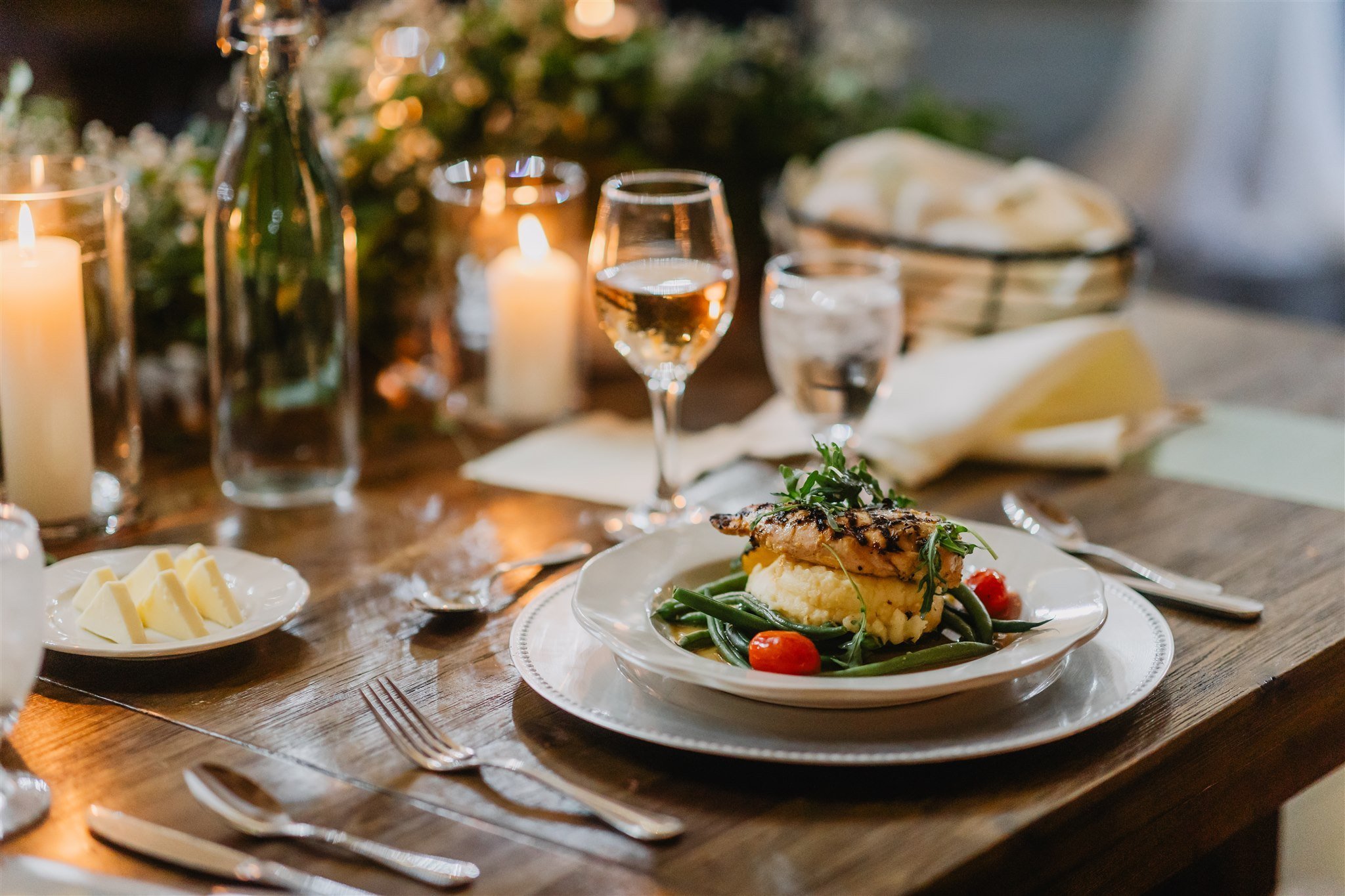 An elegant dinner plate at a wedding reception features grilled fish, mashed potatoes, green beans, and cherry tomatoes. The table is set with wine glasses, candles, and flowers.