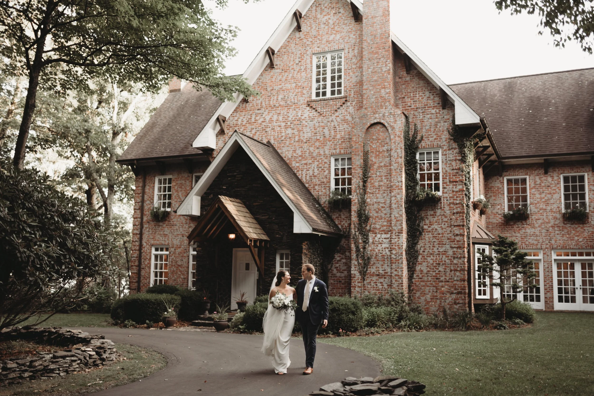 Bride and groom walking together in front of a brick estate wedding venue holding a bouquet