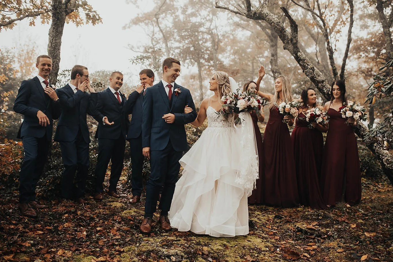 Wedding party posing and laughing in the woods with the bride and groom in the center.