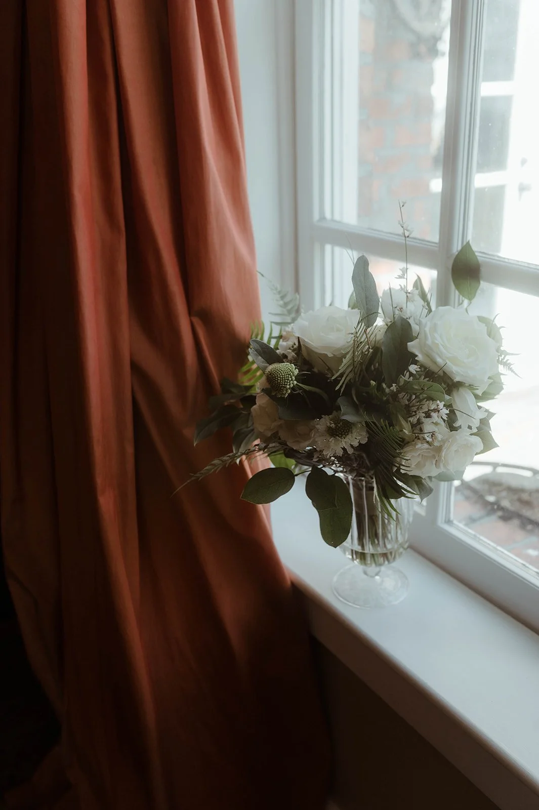 White bridal bouquet in a glass vase on a window sill beside rust-colored curtains.