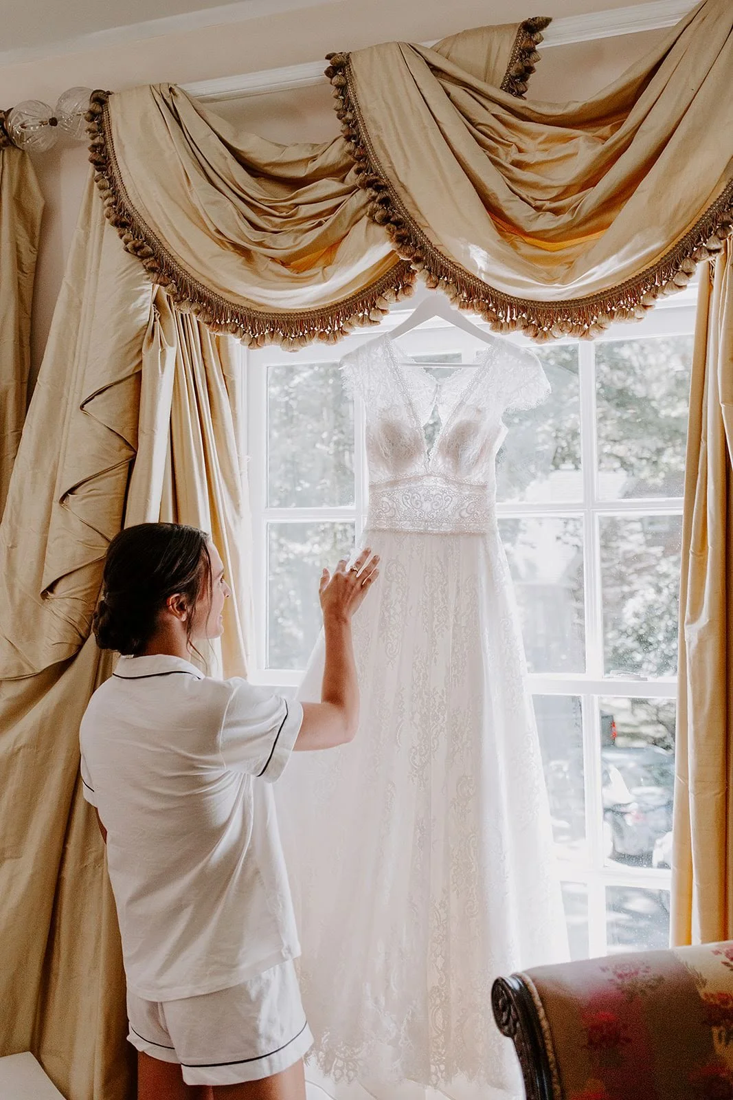 Bride admiring her wedding dress hanging in a window with elegant draped curtains.