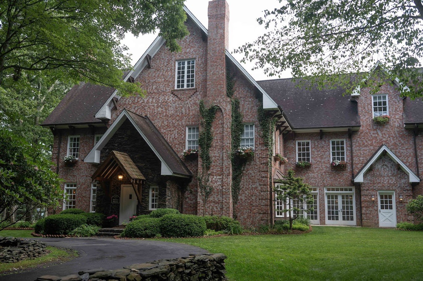 Wide view of a brick estate with steep rooflines and ivy, surrounded by trees and a curved driveway.