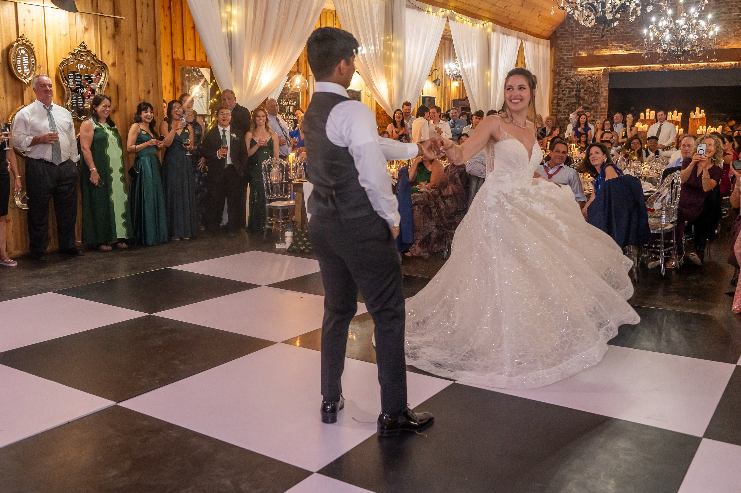 Bride twirls in a white gown during first dance as guests watch around a black-and-white dance floor in a wood hall.