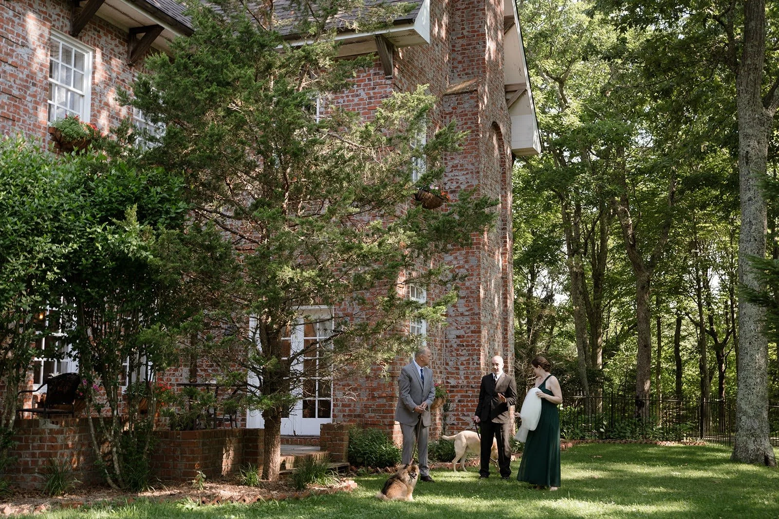 Intimate outdoor wedding ceremony beside a brick estate surrounded by trees and garden greenery.