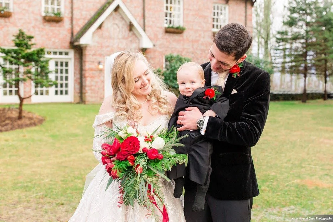 Bride and groom holding a baby outdoors in front of a brick wedding venue, all dressed in formalwear.