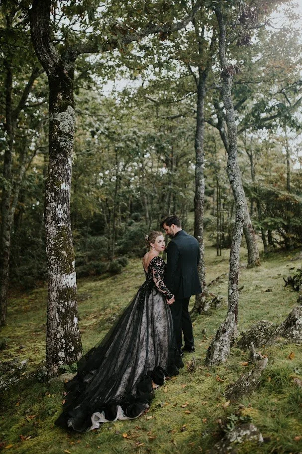 Bride and groom holding hands in a forest, showing the dramatic train of the black wedding dress.