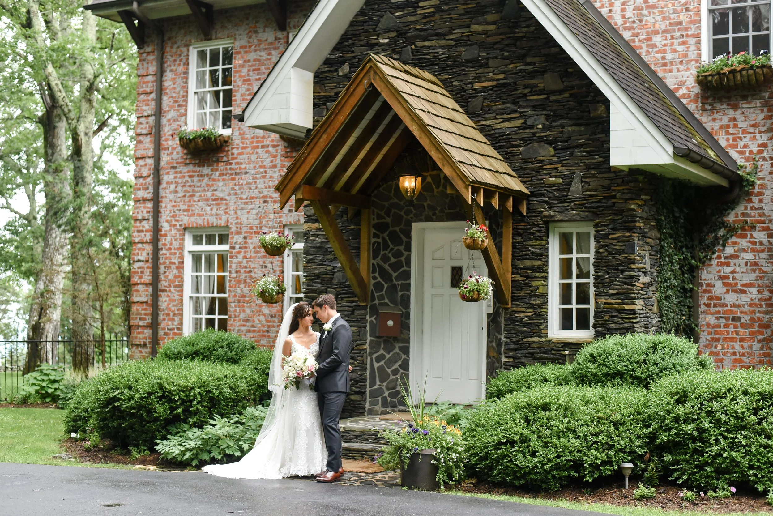 Bride and groom kissing in front of a brick and stone cottage entrance with greenery and hanging flower baskets.