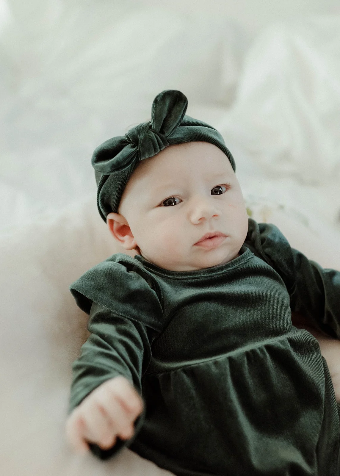 Baby wearing a dark green dress and matching bow headband lying on a soft white blanket.
