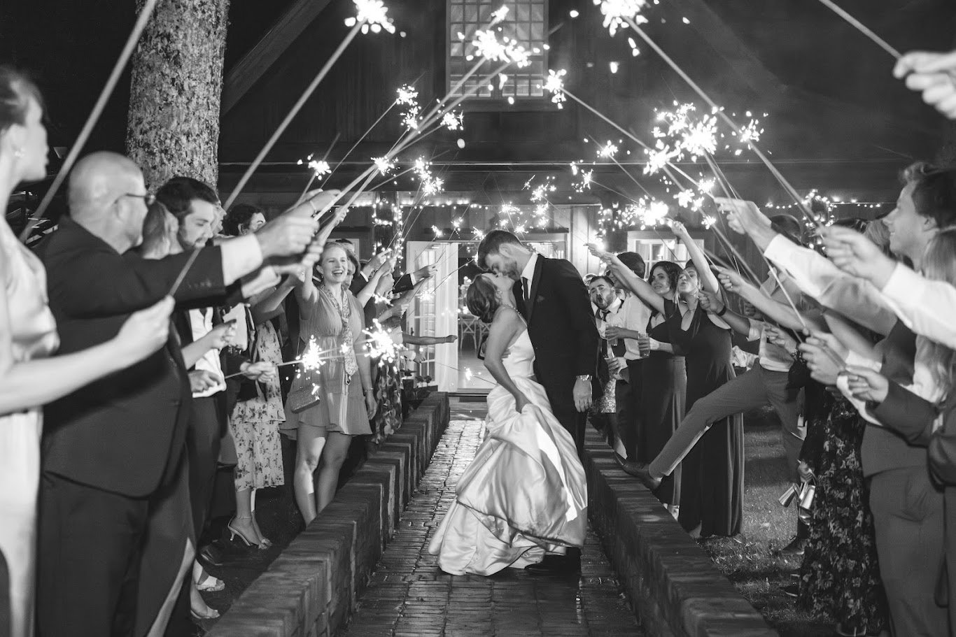 Black-and-white sparkler exit with wedding guests cheering as the bride and groom kiss at night