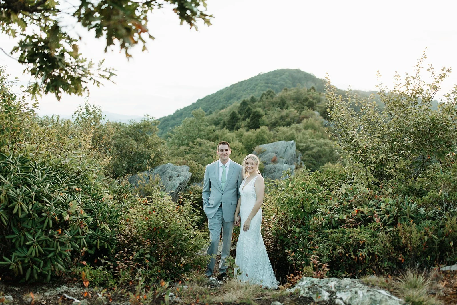 Bride and groom standing together in a lush garden setting with rocks, greenery, and mountain views behind them.