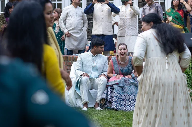Wedding guests sit on a lawn during an outdoor celebration, wearing colorful South Asian outfits.