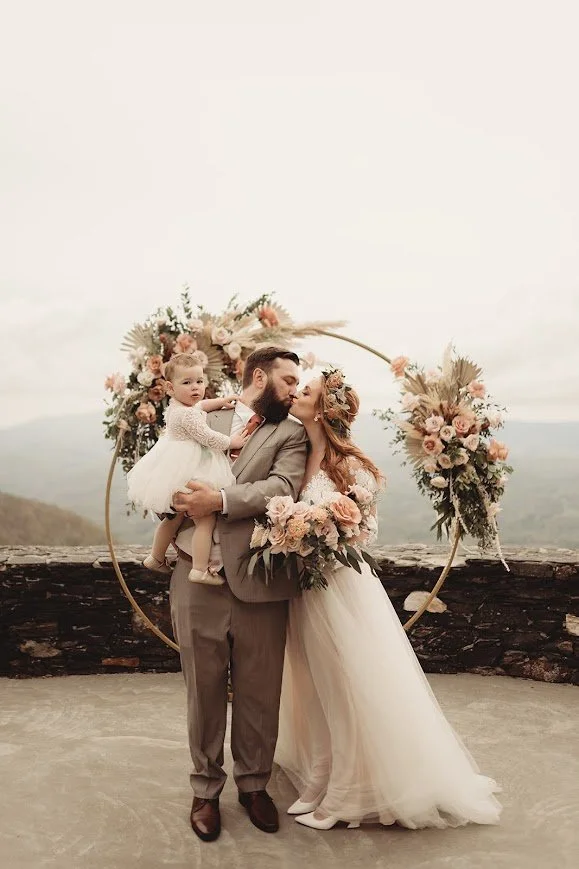 Bride and groom embrace beneath a floral ceremony arch on a stone terrace with mountain views.