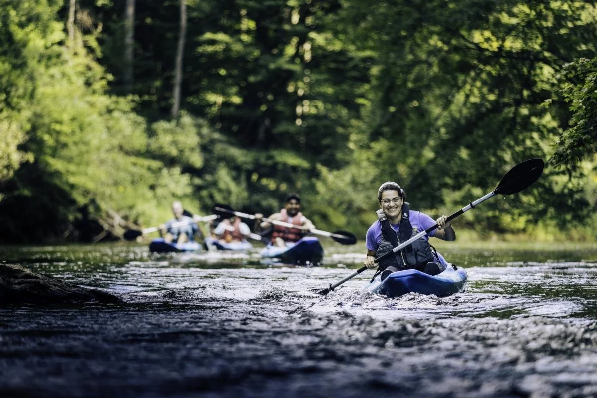 Kayaker paddling down a shallow river with other paddlers in the background