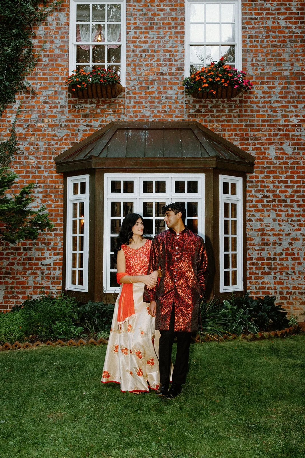 Indian couple standing together on the lawn in front of a brick estate with a bay window and flower boxes