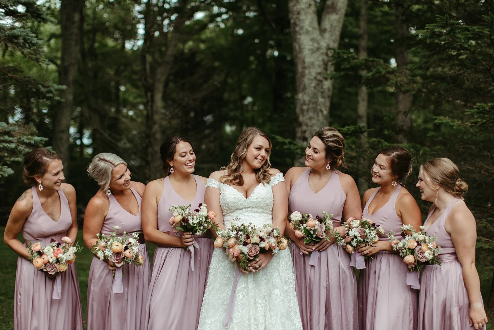Bride standing with bridesmaids in lavender dresses holding bouquets during forest wedding portraits.