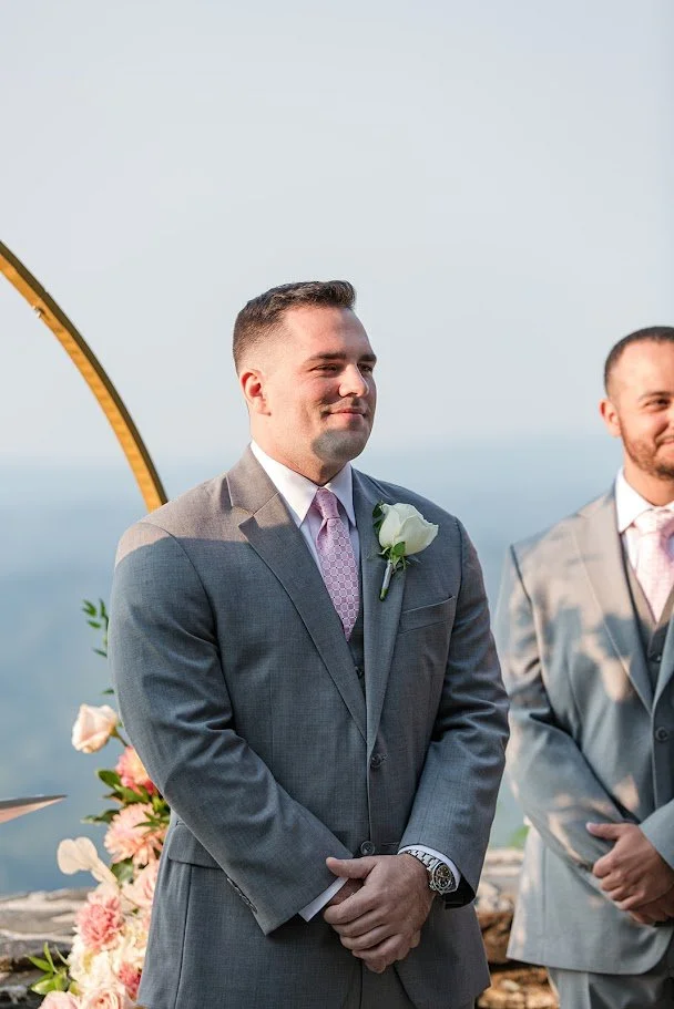 Groom smiling in a gray suit with a pink tie during an outdoor mountain wedding ceremony