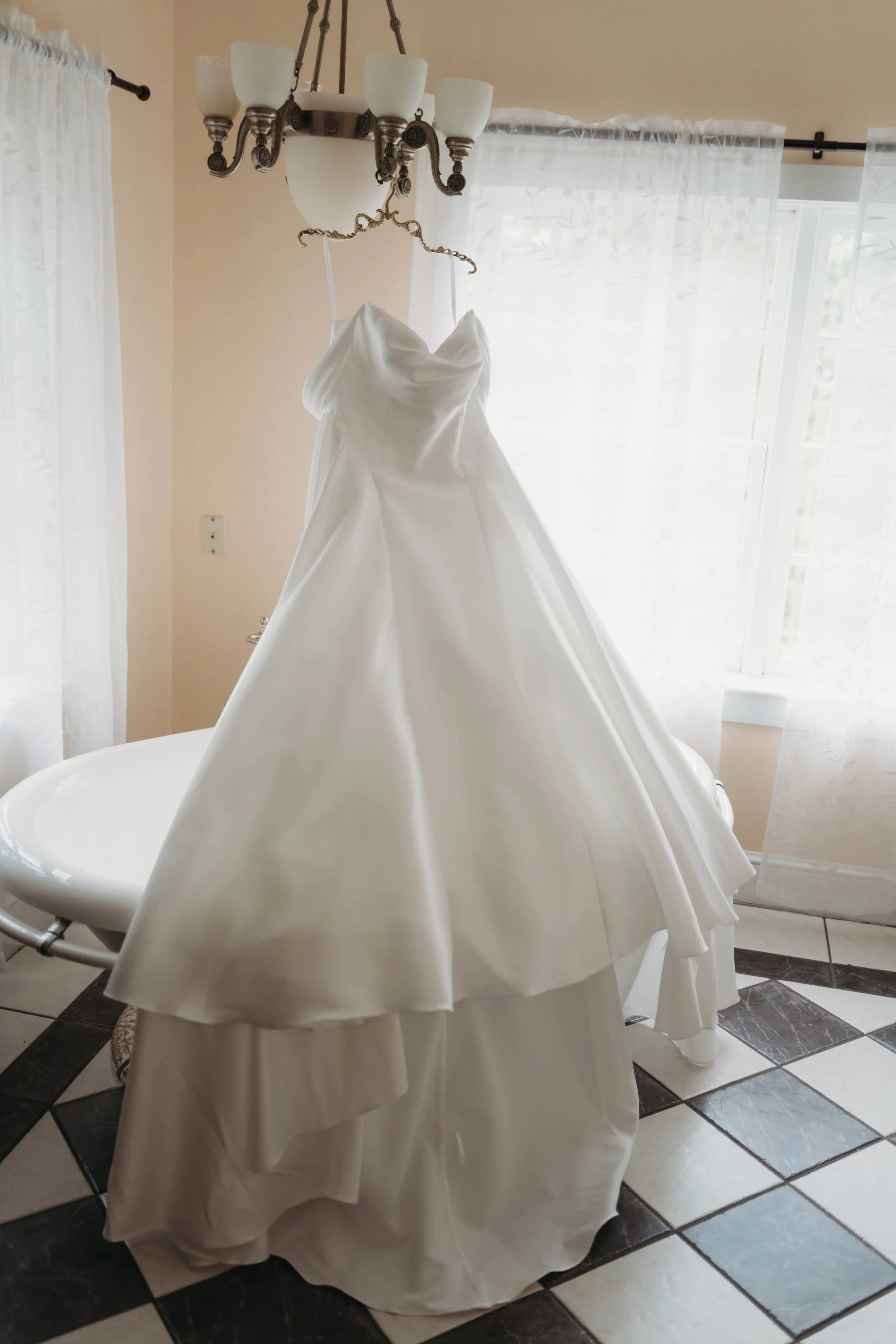 Wedding dress hanging in a bright room with large windows and black-and-white tile floor.