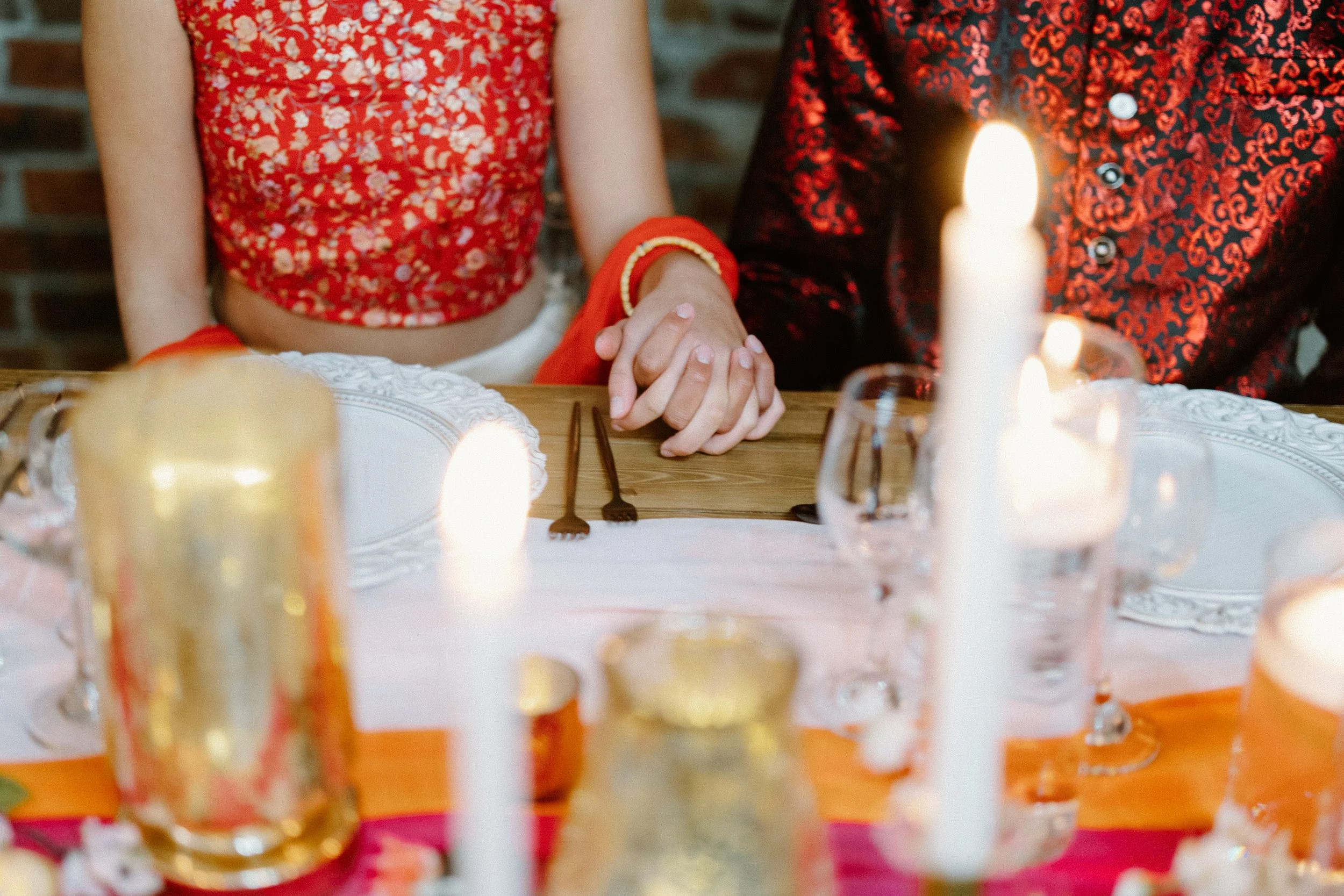 Close-up of the couple holding hands across a reception table, with glowing candles and place settings in soft focus.