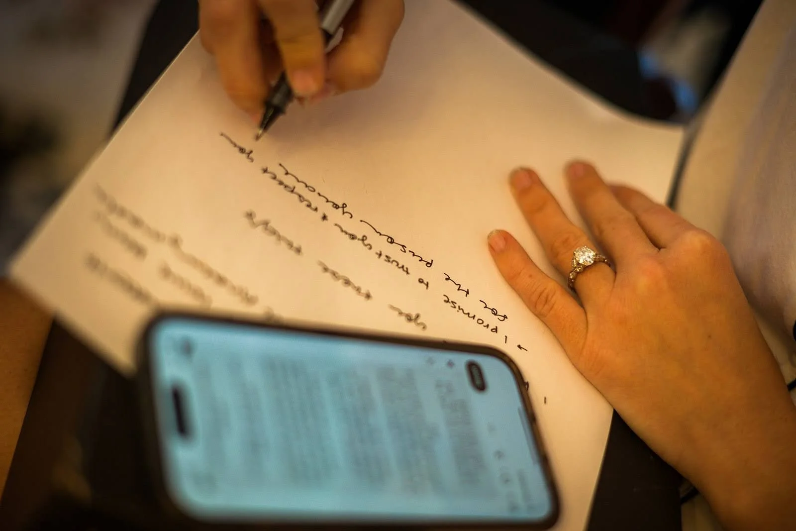 Close-up of a hand with an engagement ring writing notes on paper beside a smartphone.