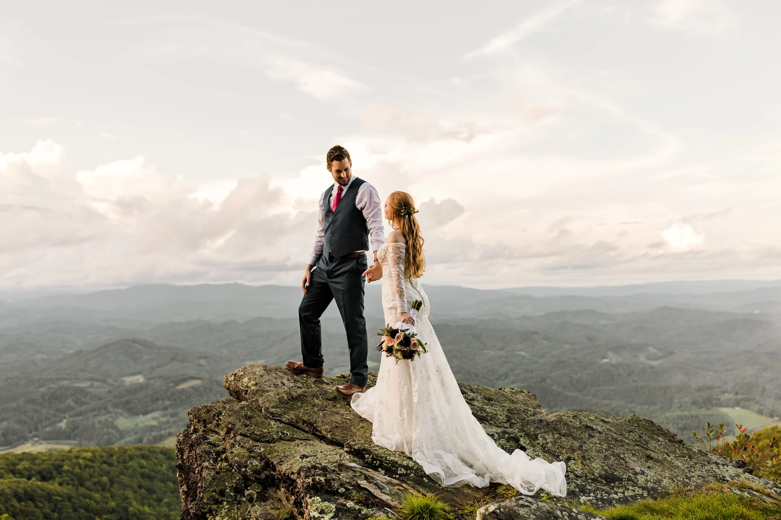 Bride and groom holding hands on a rocky overlook with mountains stretching into the distance.