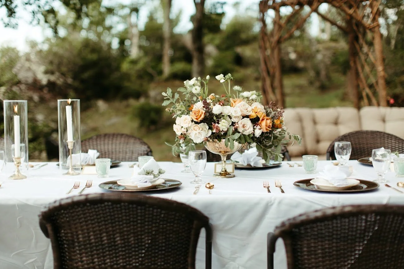 Outdoor reception table with a white tablecloth and lush floral centerpiece in a garden setting.