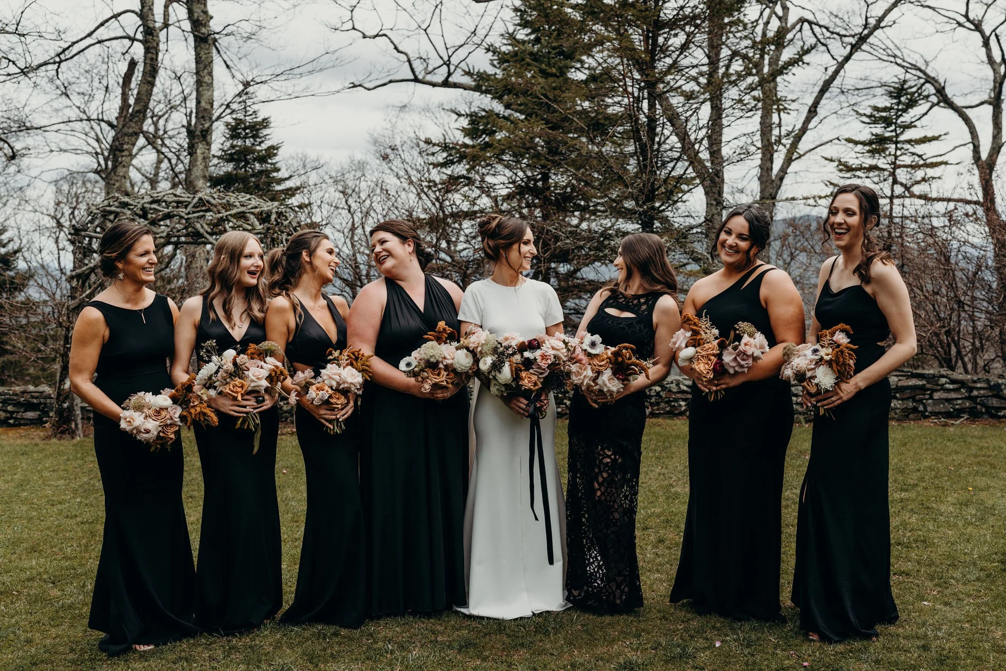 Bride with bridesmaids in black dresses holding bouquets during outdoor portraits.