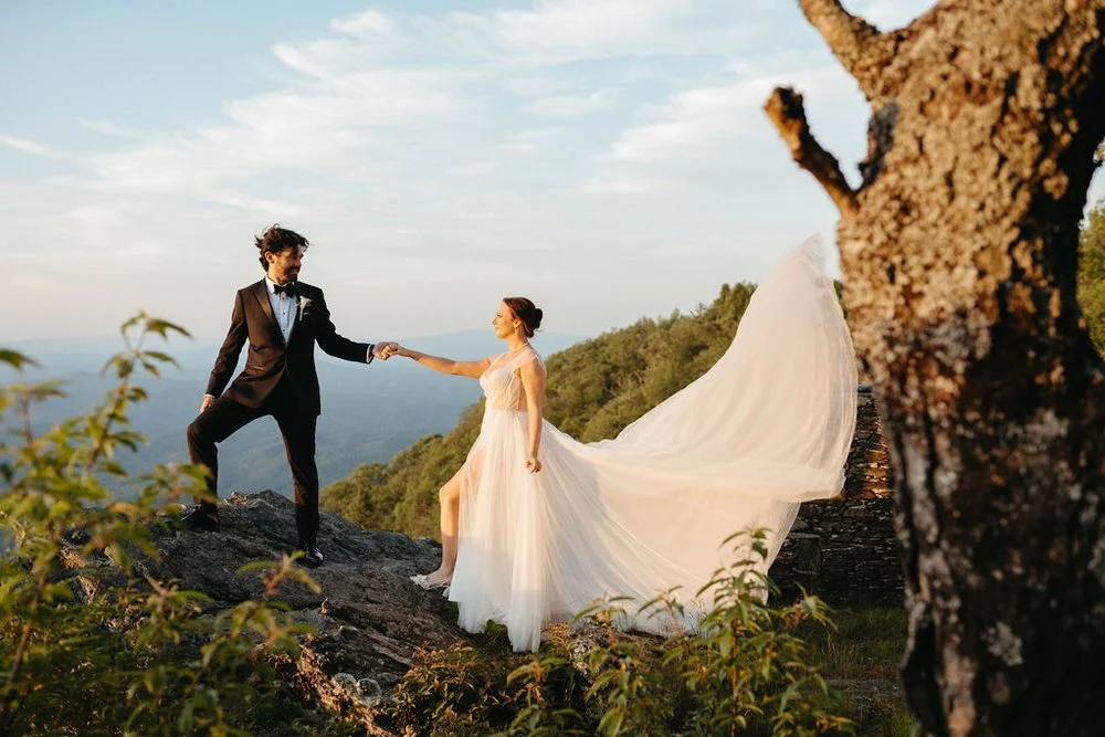 A bride in a flowing white dress and a groom in a black tuxedo hold hands on a rocky cliff at sunset, overlooking a scenic mountain vista under a blue and cloudy sky. A large tree trunk is visible in the foreground.