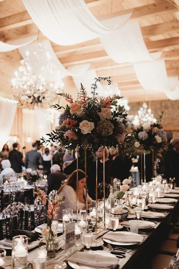 Romantic reception table with tall floral centerpieces, chandeliers, and soft white draping overhead in a glowing venue.