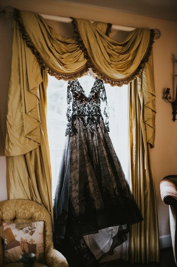 Black lace wedding dress hanging in front of a bright window framed by gold drapes.