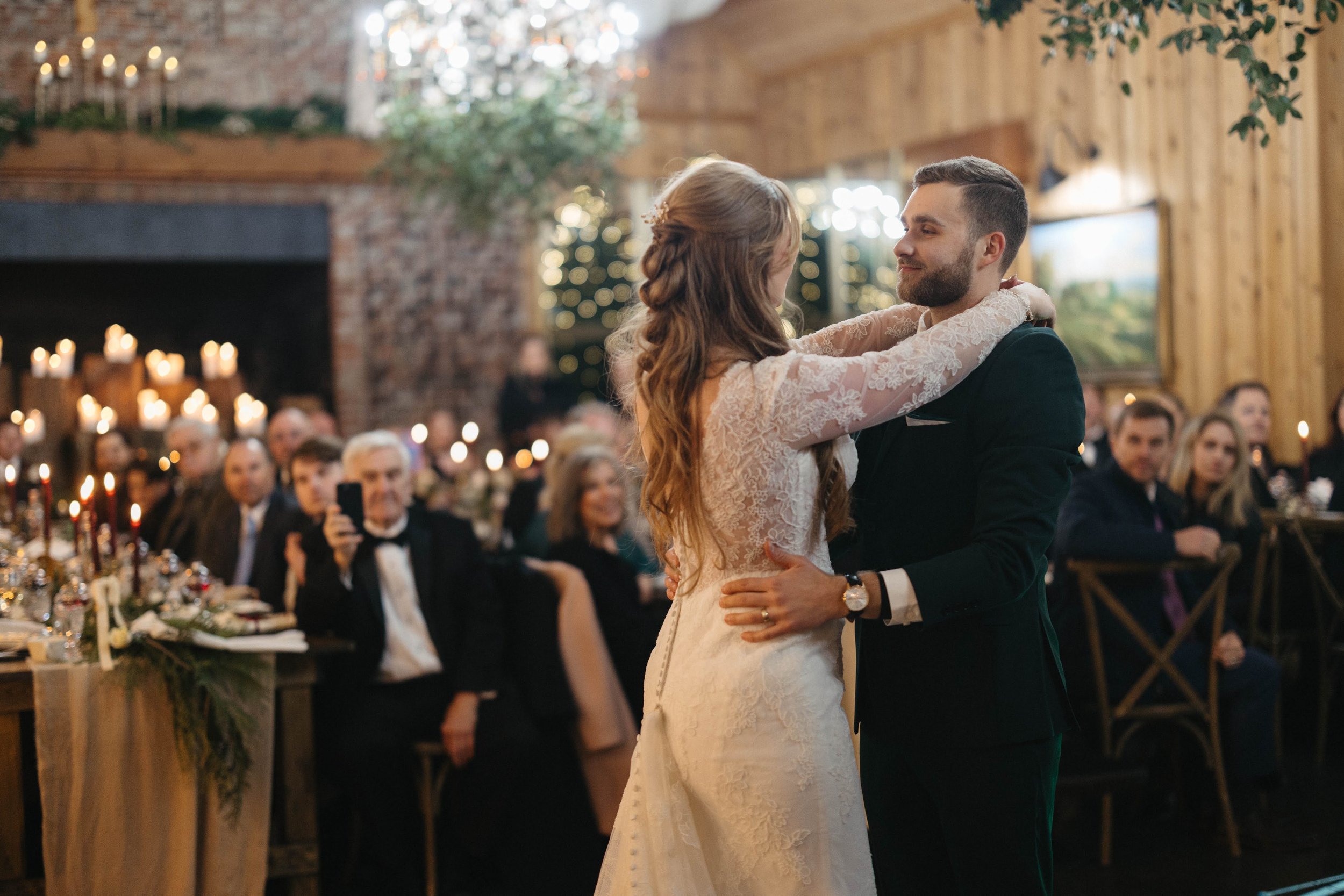 Bride and groom share their first dance in a candlelit reception room while guests watch from their tables.