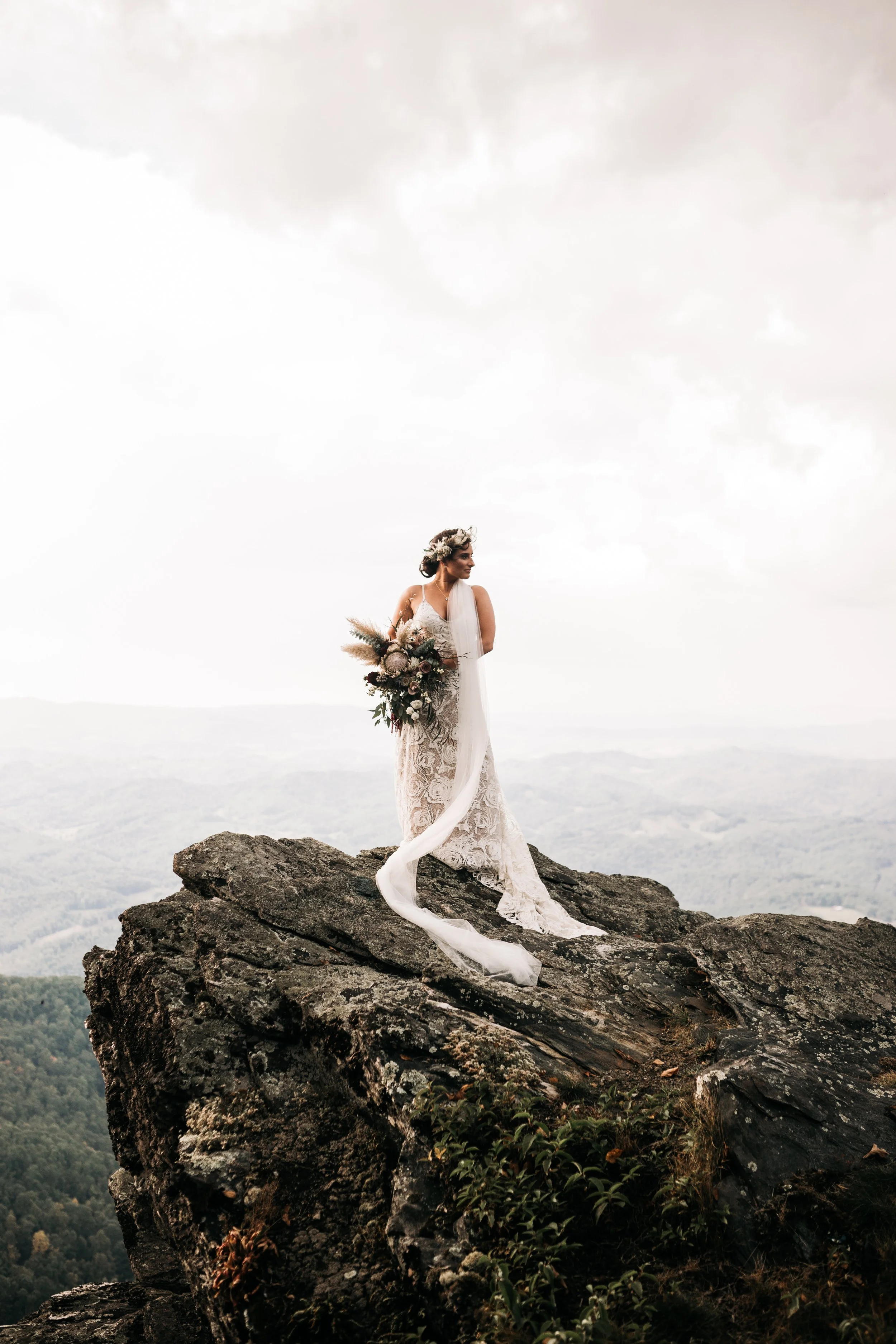 Bride stands on a rocky cliff holding a bouquet, with a long veil trailing behind and mountains in the distance.